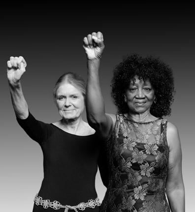 Gloria Steinem and Dorothy Pitman Hughes stand next to each other, each raising one arm with a closed fist.