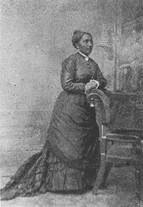 A full-length black and white studio portrait of Black woman. She wears a long dress with overskirt, bustle, and lace, and a neck brooch and earrings.    