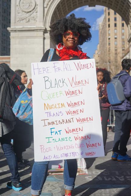 Woman holding a sign that reads: Here For All Women Black Women, Queer Women, Muslim Women, Trans Women, Immigrant Women, Fat Women, Islamic Women, Poor & Working Class Women, and all other types of Women.
