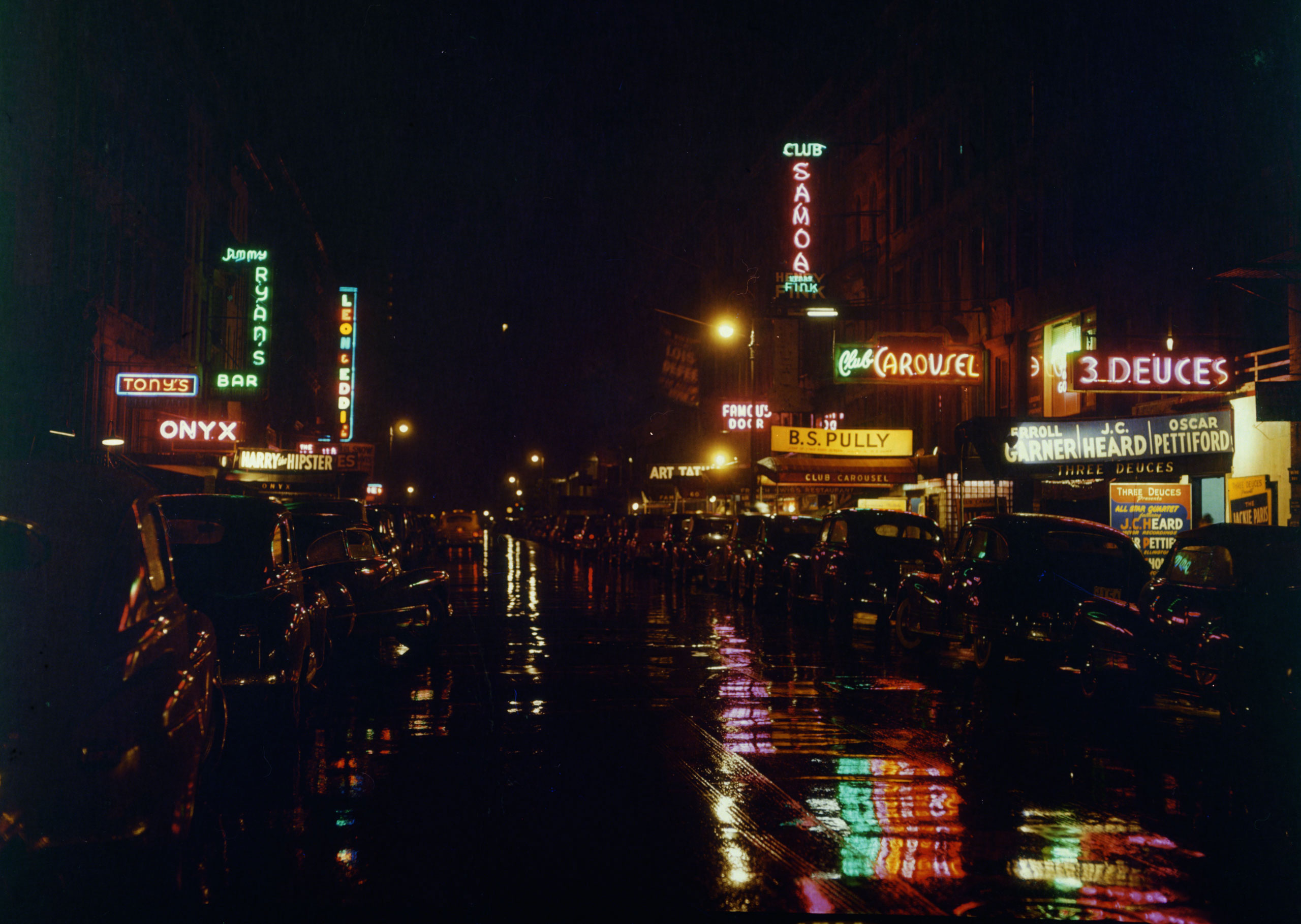Road at nighttime, with neon signs reflecting on the wet pavement, and parked cars on either side of the street