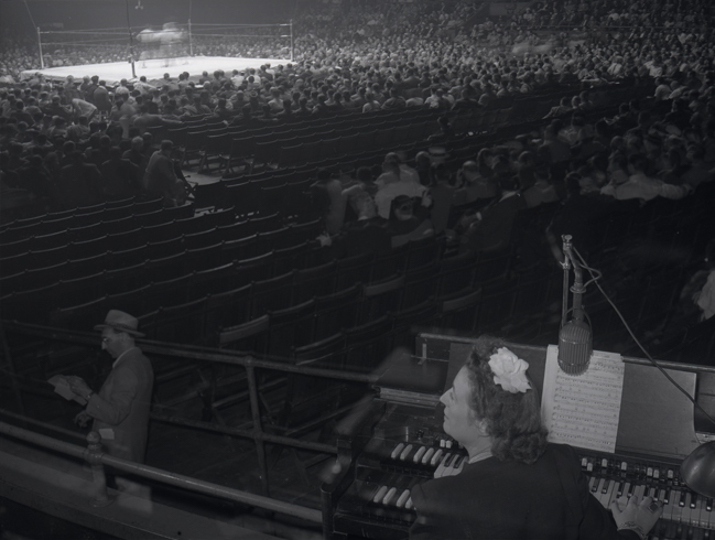 Sports organist Gladys Gooding sits at an organ at Madison Square Garden with a boxing ring and audience in the background.