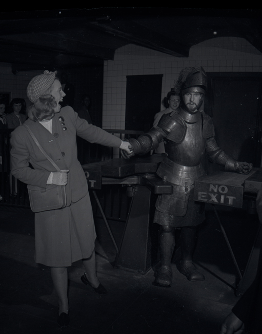 A man in metal armor walks through a subway turnstile while a woman in a suit holds his hand and leads him through.