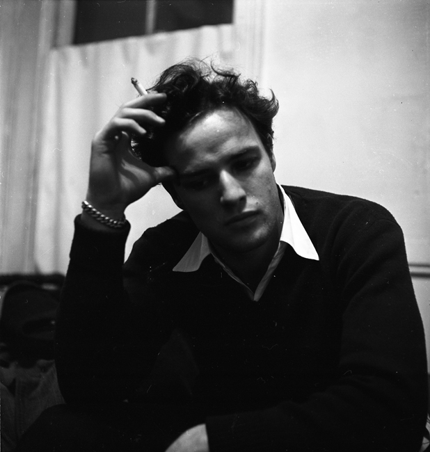 Actor Marlon Brando sits indoors, wearing a sweater and holding a cigarette.