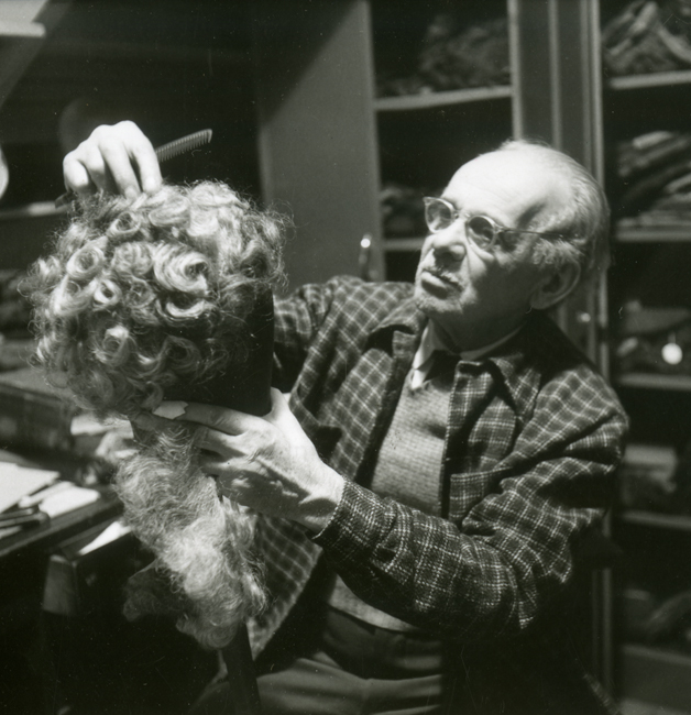 A man wearing glasses holds a long curly wig belonging to the Metropolitan Opera out in front of him.