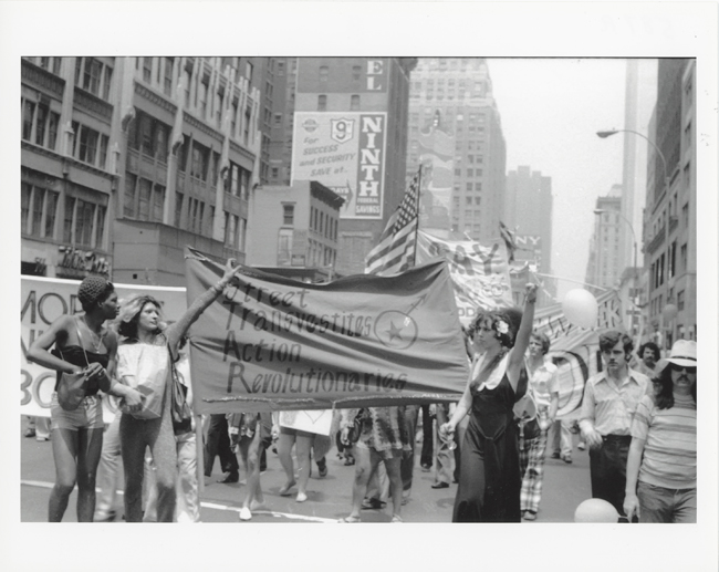 Black and white photograph featuring a parade during Christopher Street Liberation Day. Three figures, in front hold a banner for STAR, or Street Transvestite Action Revolutionaries.
