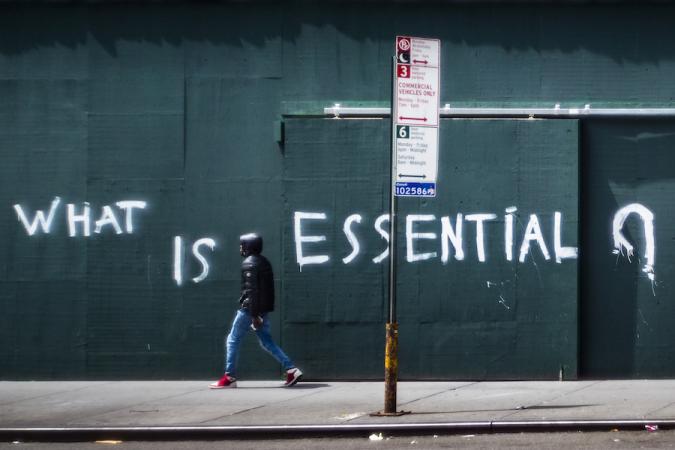 A man walks by a boarded up wall with the words "What is essential" spray painted across it.