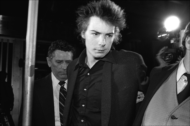 Black and white photograph of Sid Vicious. A man to his left grabs his arm and leads him away.