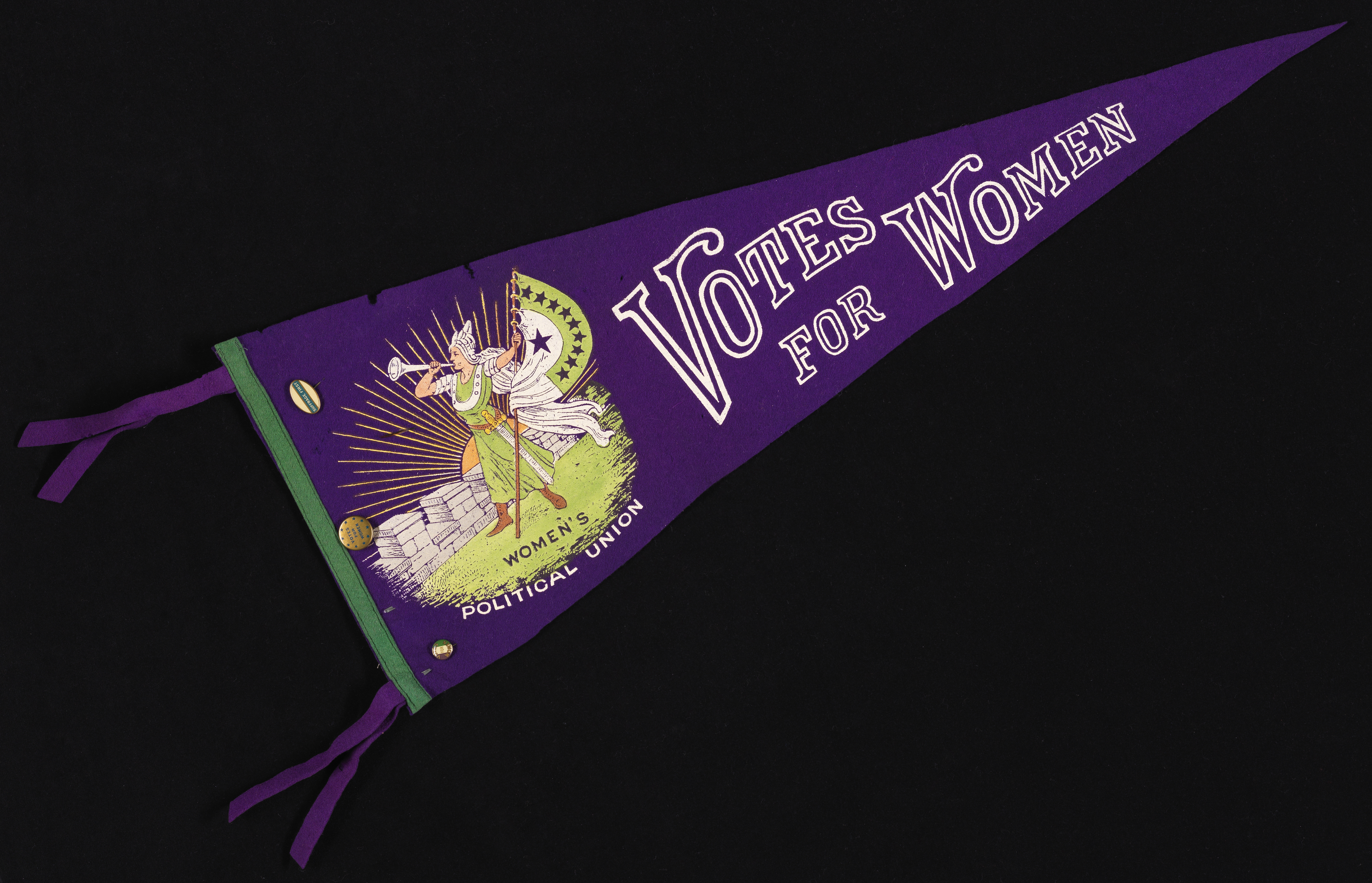 A photograph of a wool purple pennant on a black background. White text on the pennant boldly says, “Votes for Women,” fixed next to an image of a women blowing a horn and holding a flag.