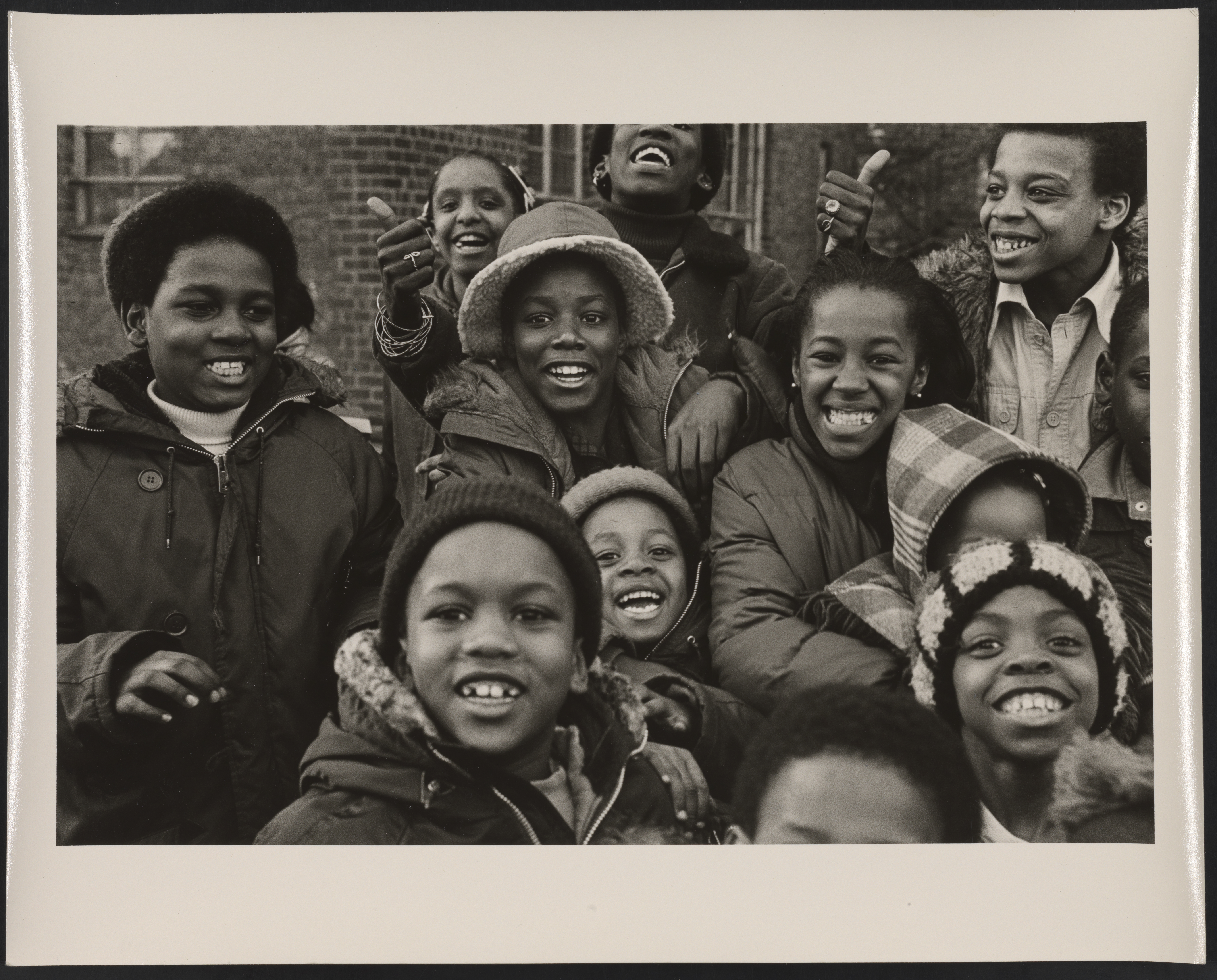 Black and white photograph of a group of Black-presenting children wearing winter clothes and smiling at the camera