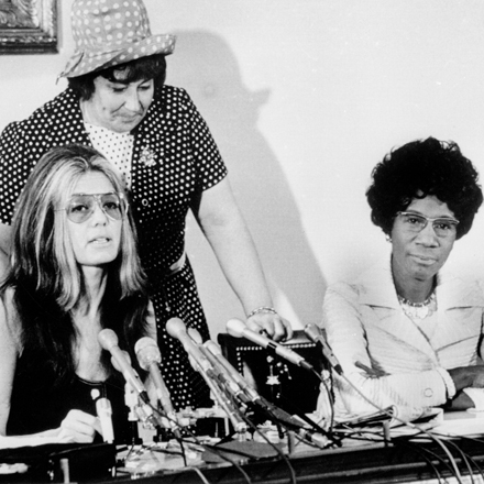 Three women sit at a table, one has microphones in front of her, suggesting it is a press conference 