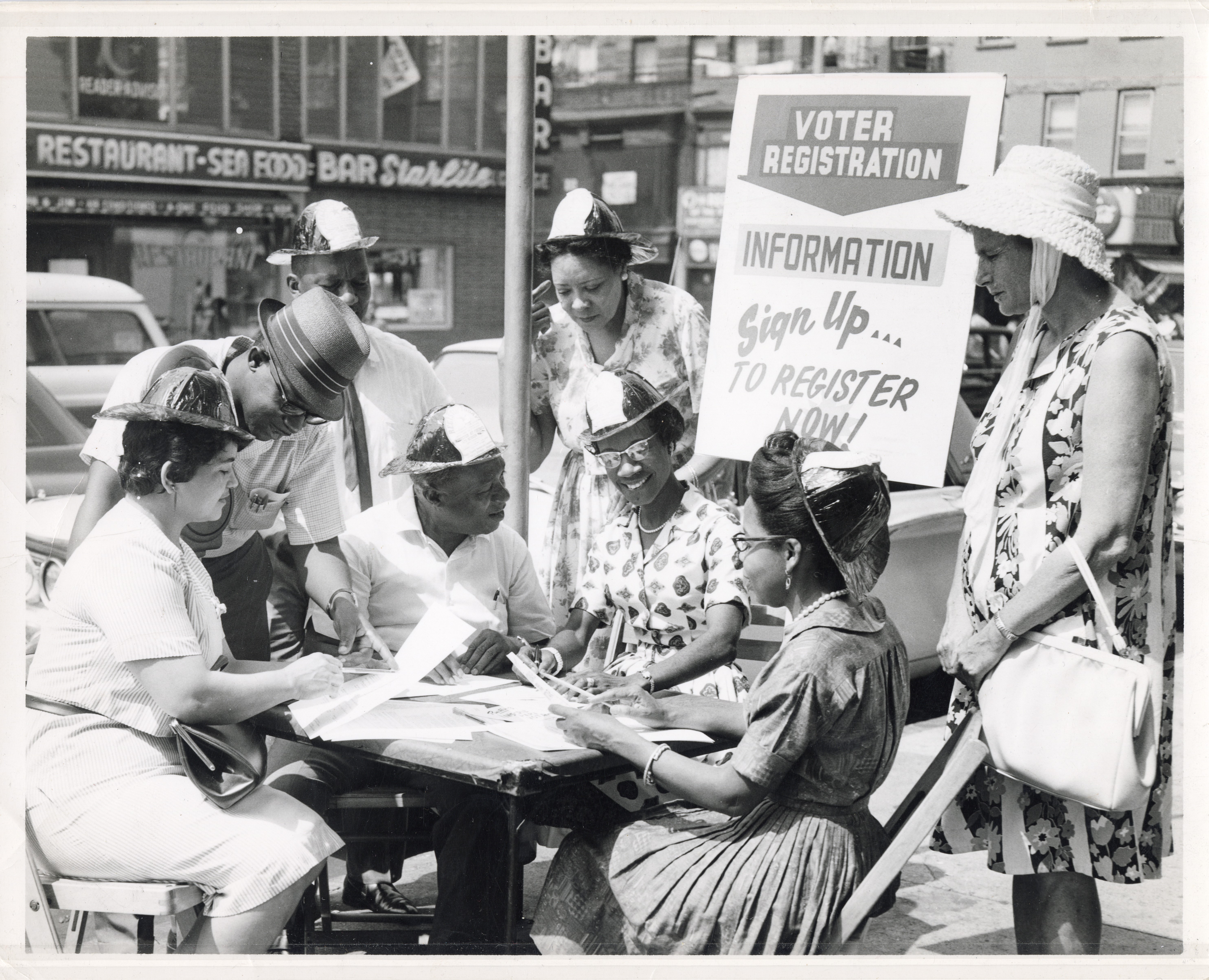 A black and white photograph of eight people sitting around a table on a city sidewalk. In the middle of the group is Shirley Chisholm, sitting under a sign saying, “Voter Registration Information.