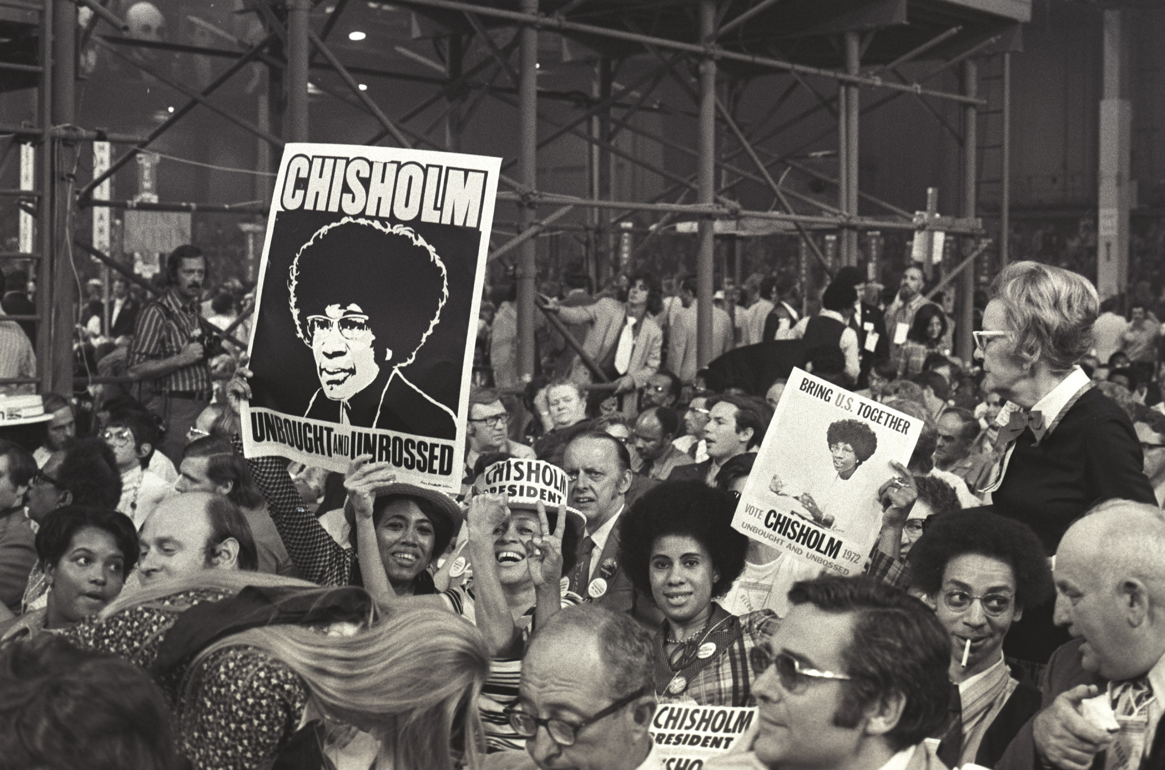 A black and white photograph of a crowd gathered in support of candidate Shirley Chisholm. Several people are smiling and holding up signs supporting her campaign for president.