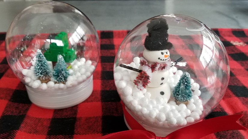 Image of two snow globe crafts. A winter scene with evergreen trees to the left, and a snowman and tree to the right.