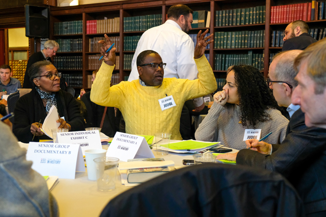 A group of History Day judges sit at a table. A man with a yellow sweater has his arms raised, mid-discussion.
