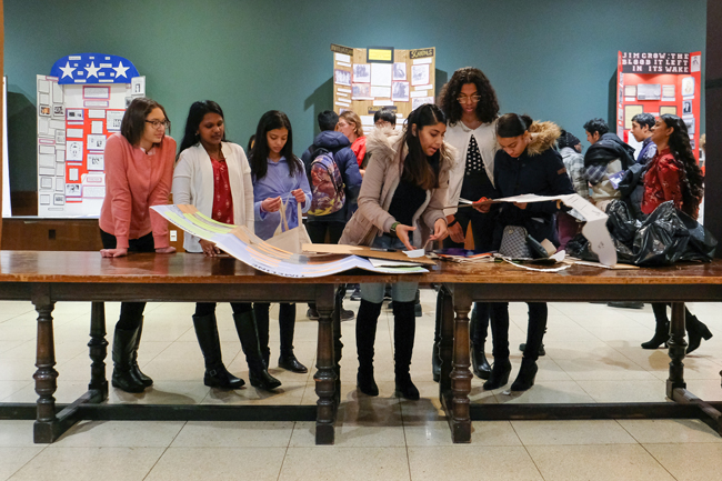 A group of students stand on one side of a long wooden table looking at documents and History Day projects.