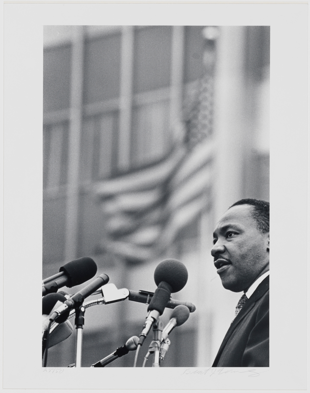 Black and white photograph of Martin Luthur King Jr. standing at a microphone with a building and flag waving behind him.