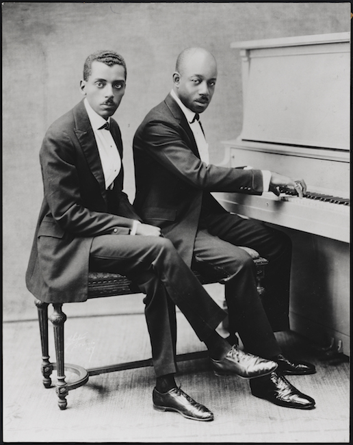 Two African-American gentlemen in suits sit on a piano bench, with a piano to the right. The man on the right has his hands on the keyboard.