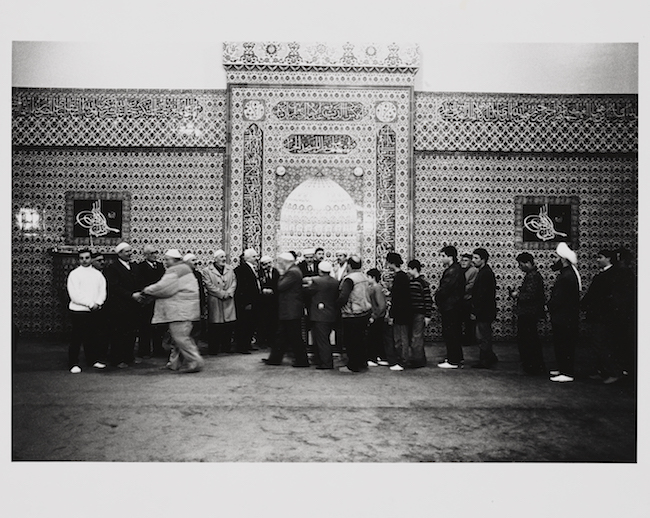 A group of Muslim men of various ages, are gathered outside of a Mosque in Brooklyn. They are greeting each other and standing in line.
