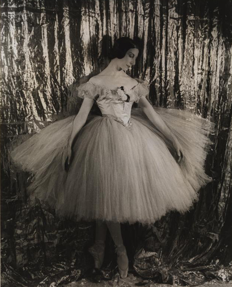 A portrait of a ballerina, in costume, standing en pointe in front of a curtain.