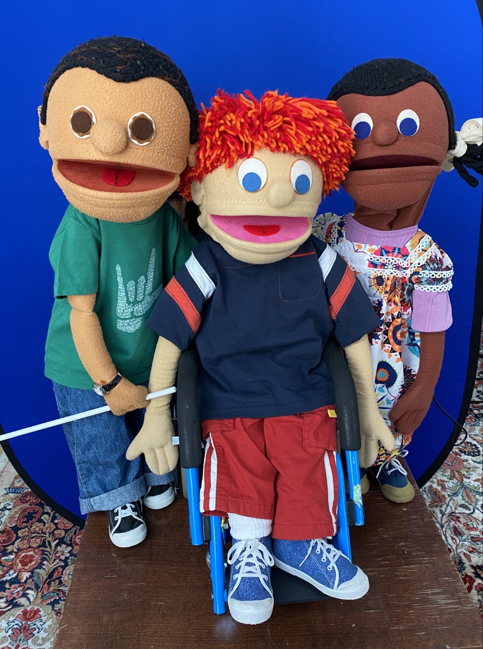 3 puppets created by NYC Kids Project standing on brown wood table, with bright blue background. One male puppet, Latinx, with dark hair, green tshirt , blue jeans , is holding a white cane. One male puppet with light skin and red hair uses a wheelchair and wears red pants, blue shirt with stripes on the arms. One female puppet, African-American has a colorful flowered dress over a lavender shirt and hair in two braids with white bows.  