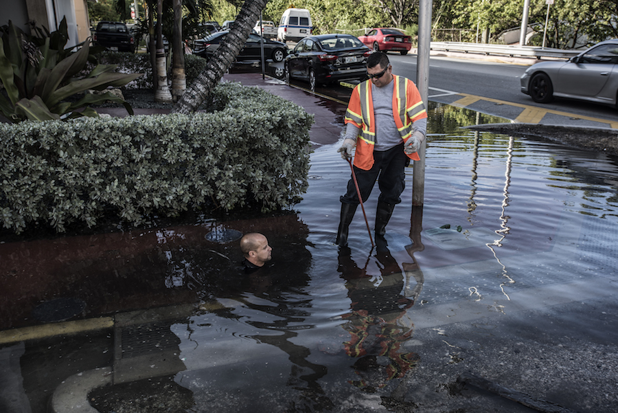 A man's head is visible popping out of the water in a flooded intersection. Next to him, a man stands with water to his ankles.