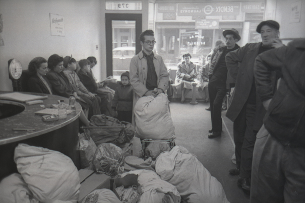 Several men and women stand and sit in a Greenwich Village laundromat with bags of laundry and washing machines sitting in front of them.