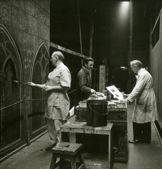 Three men in smocks and aprons hold paintbrushes and papers as they paint sets for the Metropolitan Opera with work tables and paint buckets beside them. 