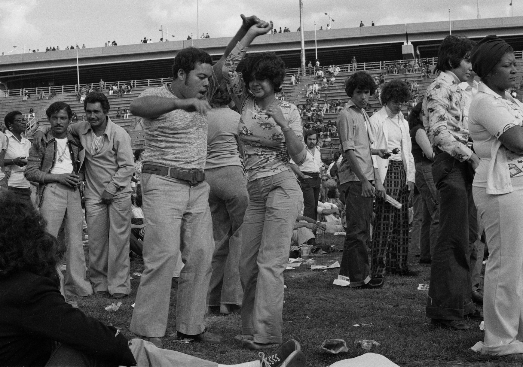 A casually dressed couple dances salsa with people in the background on Randall's Island in 1974.