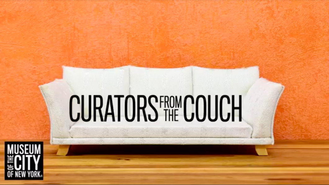 White couch against an orange painted wall and wood floor. Text on the couch reads "Curators from the Couch" in black letters.