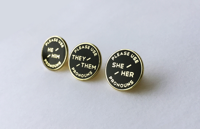 Three enameled pronoun pins. Black background with gold text. 