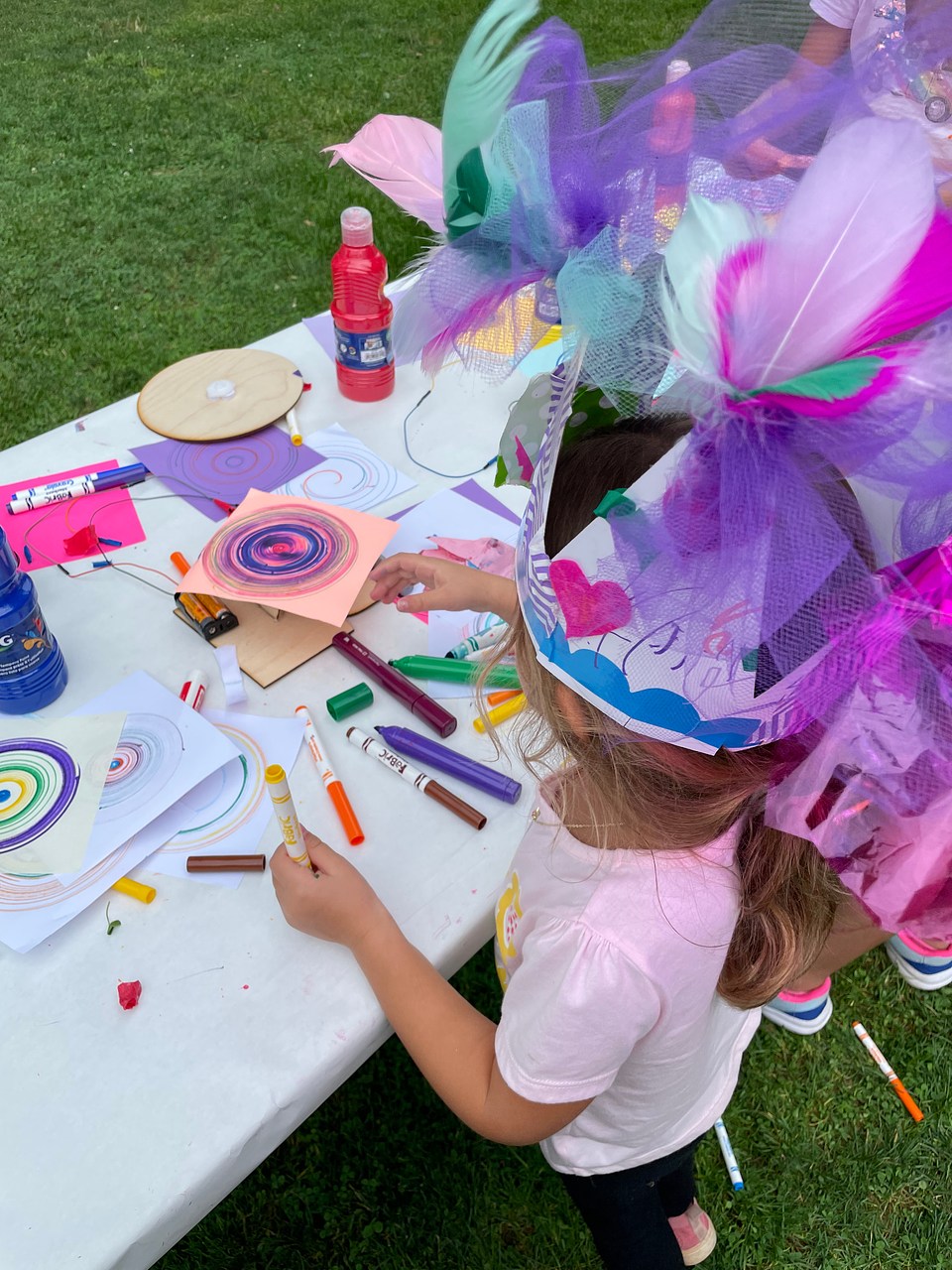 A child draws with markers while wearing a large purple hat.