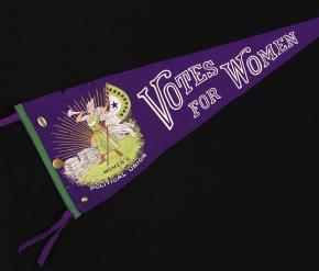 A photograph of a wool purple pennant on a black background. White text on the pennant boldly says, “Votes for Women,” fixed next to an image of a women blowing a horn and holding a flag.