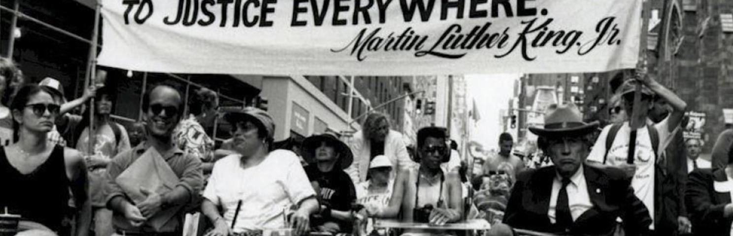 A crowd of people with disabilities and individuals in wheelchairs gather under a banner that reads "Injustice Anywhere is a Threat to Justice Everywhere" Martin Luther King Jr.