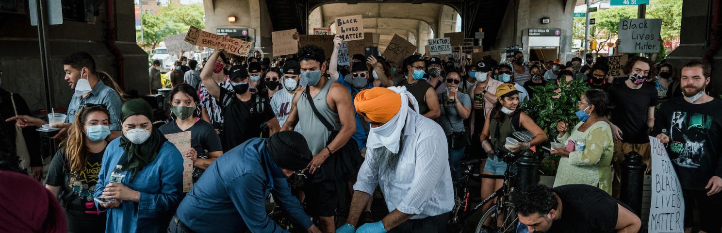 A crowd of people stand around a table with food containers. The man in the center is opening a case of water bottles to distribute.