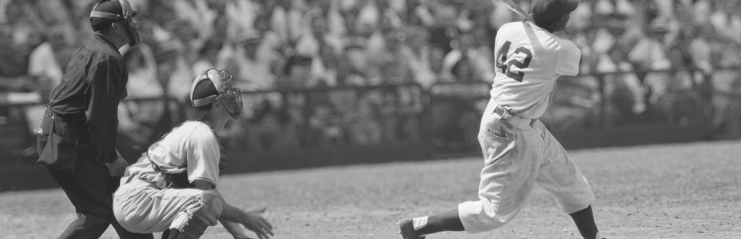 Jackie Robinson stands ready to bat in a crowded stadium as the pitcher and umpire look on from behind Robinson