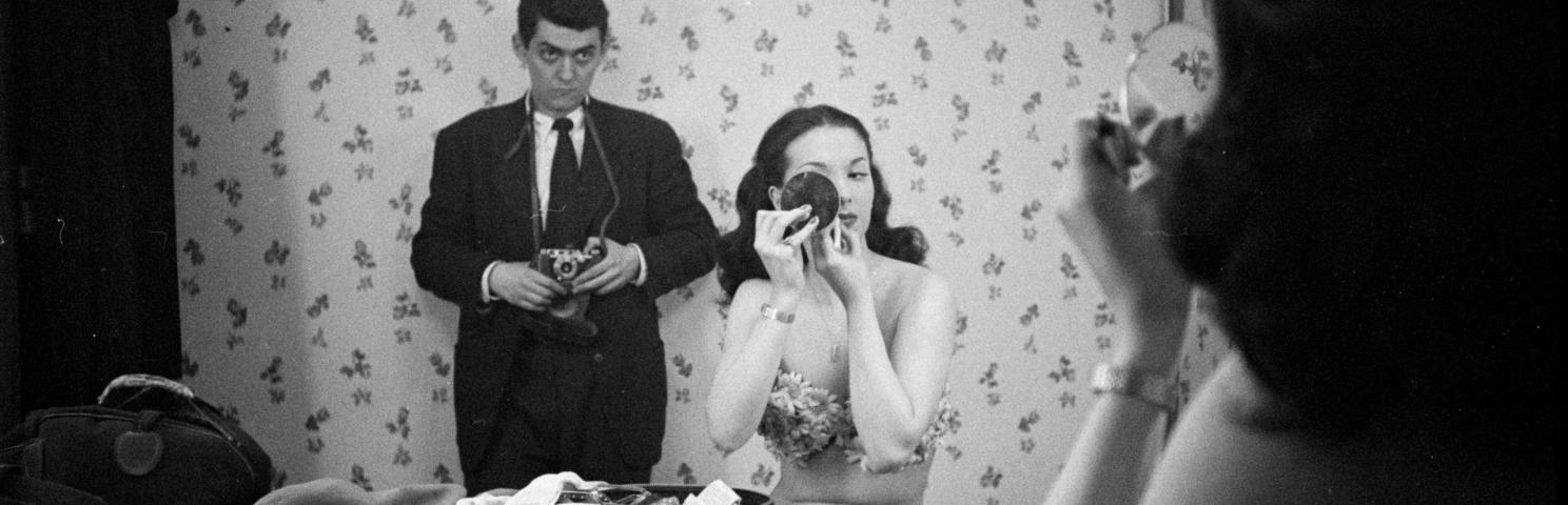 Taken in a mirror’s reflection, a woman uses a compact mirror to apply makeup, while photographer stands behind her watching