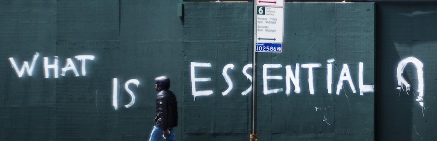 A man walks by a boarded up wall with the words "What is essential" spray painted across it.