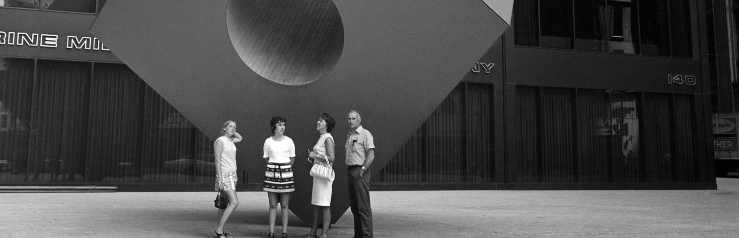 Four people stand on the sidewalk in front of a large statue of a cube with a hole going through the center of it