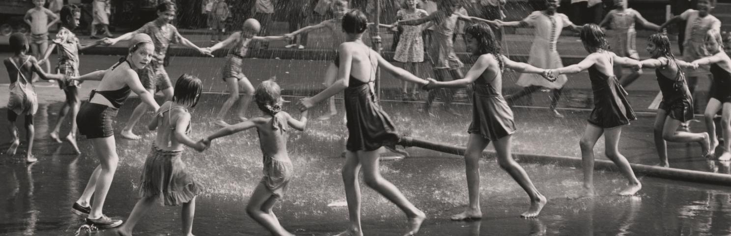 A group of children and teenagers hold hands in a circle as they run around a playground sprinkler