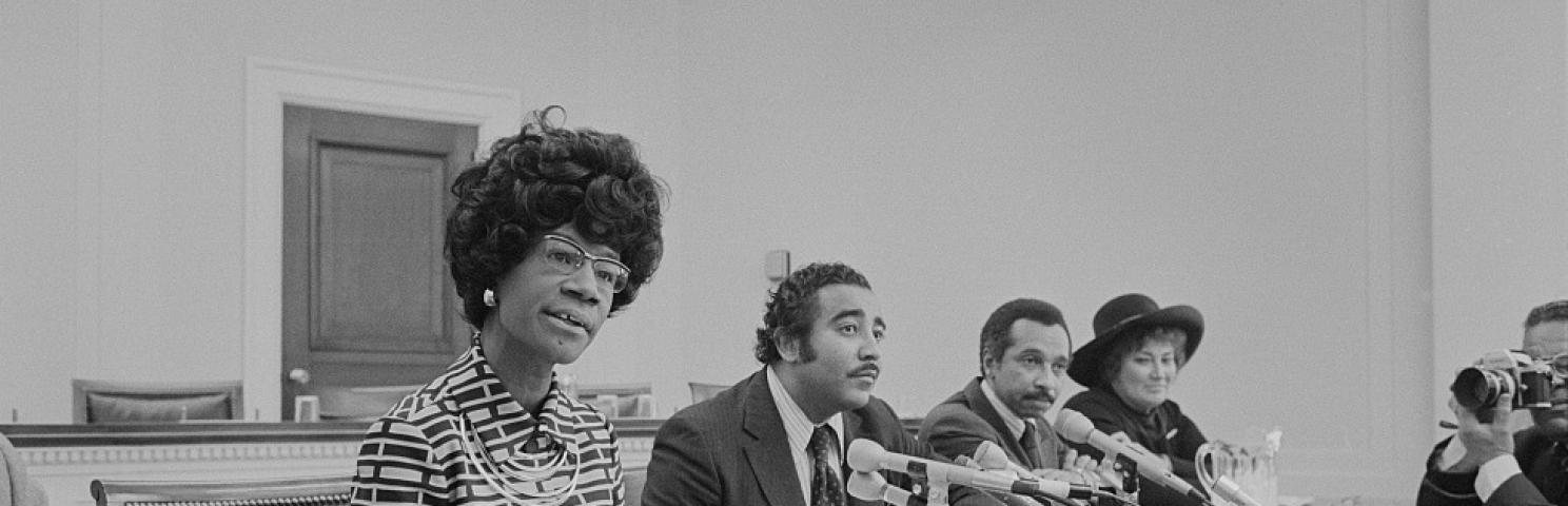 Photograph shows Rep. Shirley Chisholm, Rep. Parren Mitchell, Rep. Charles Rangel, and Rep. Bella Abzug seated at a table with microphones