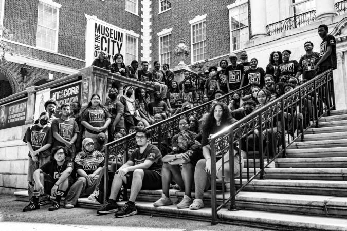 Black and white group photo of about 70 people wearing Museum of the City of New York shirts standing and sitting on the Museums Fifth Avenue entrance steps.