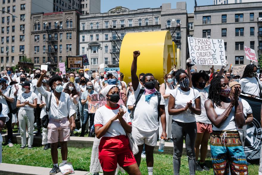 A crowd of protestors who are racially mixed but predominantly Black stand in solidarity in the plaza outside of the Brooklyn Museum. Some are holding signs, others are raising fists. All are wearing white. 