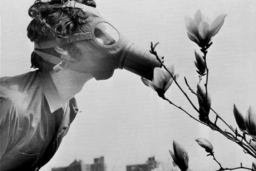 Black and white photograph of a man wearing a gas mask leaning over to smell flowers. In the background is a city skyline. 