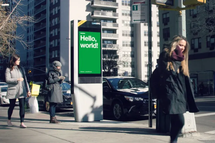 People walk down the street in front of a LinkNYC screen.