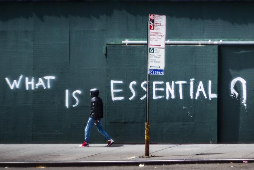 A man walks by a boarded up wall with the words "What is essential" spray painted across it.