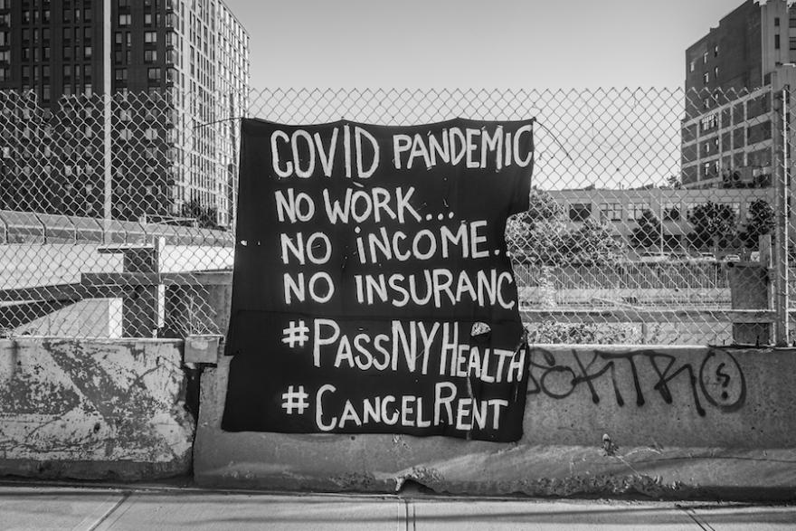 A black and white photograph of a sign on a fence that reads "COVID PANDEMIC No work....No Income...No Insurance...#PassNYHealth #CancelRent"