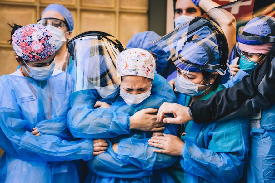 A group of medical workers stand holding each other wearing PPE including masks, face shields, and gowns.