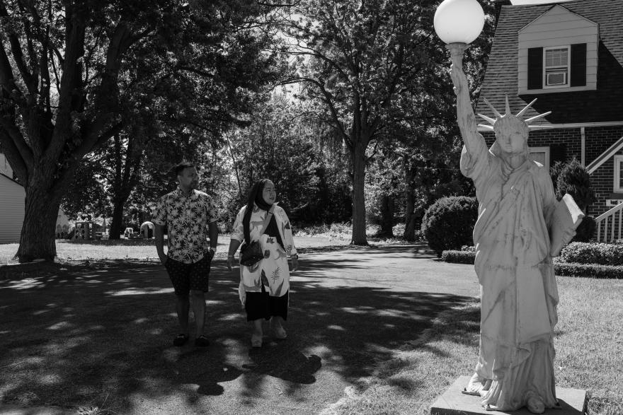 A man and a woman stand on a lawn next to a small replica of the Statue of Liberty.