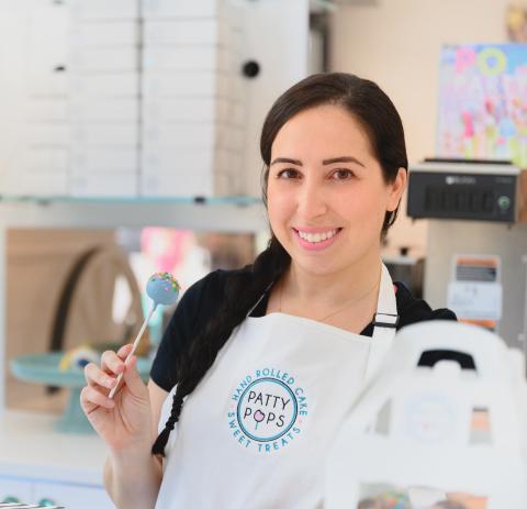 Colored photograph of woman with a sleek braided ponytail smiling while holding a cakesicle in her right hand and wearing an apron with the logo Patty Pops