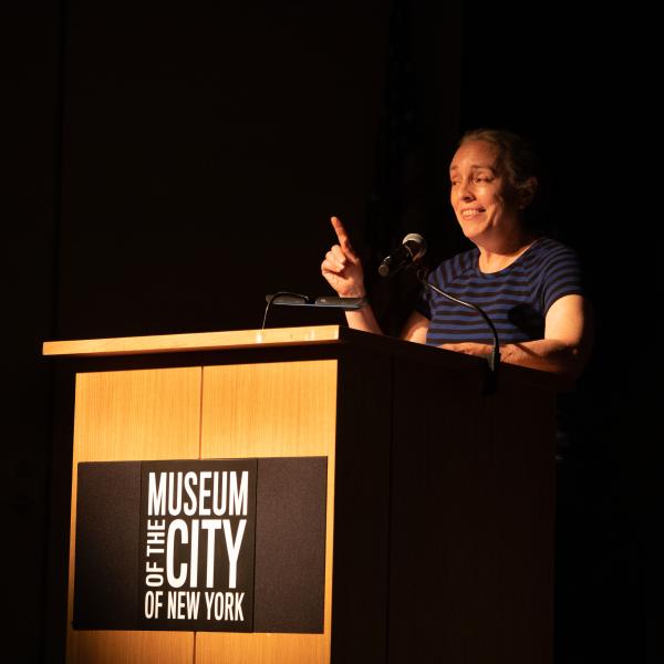 Image of woman in front of a podium labeled "Museum of the City of New York". The background is black.